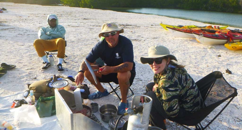 Three people sit in chairs on a white sandy beach and prepare food. 